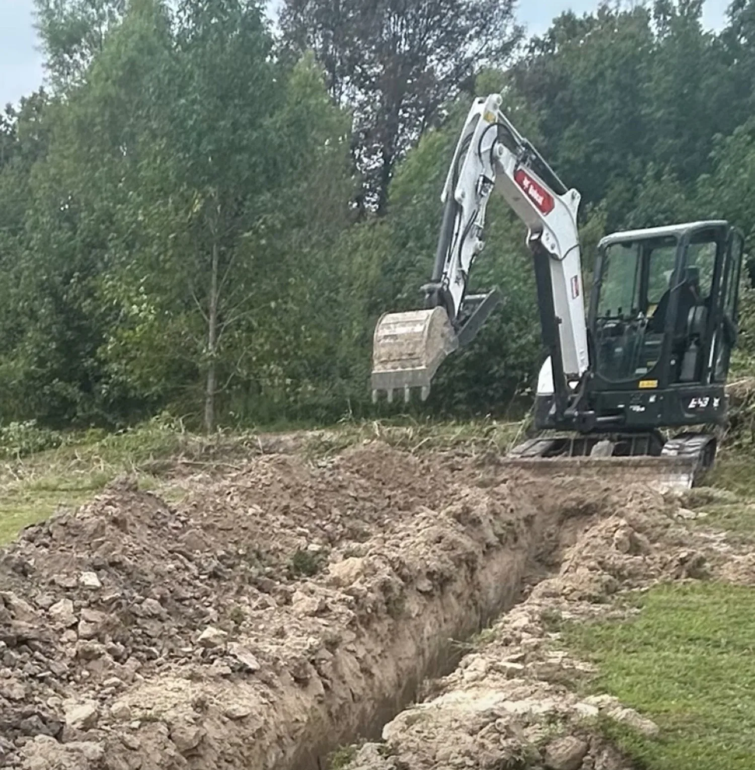 A mini excavator beside a freshly dug trench at the edge of the woods